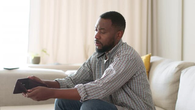 Financial problems. Young depressed unemployed african american man sitting at home with empty wallet, free space