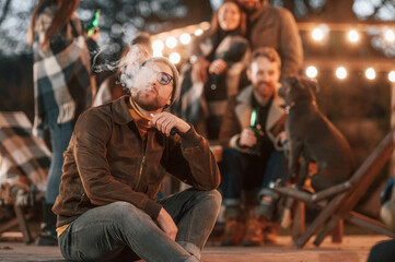 Man is smoking. Group of people is spending time together on the backyard at evening time