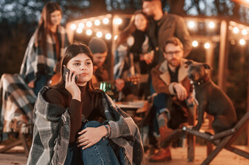 Woman with smartphone is sitting. Group of people is spending time together on the backyard at evening time