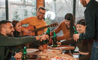 Man is playing guitar. Group of friends is having good weekend indoors in the wooden building together