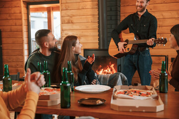 Beer, pizza and guitar. Group of friends is having good weekend indoors in the wooden building together