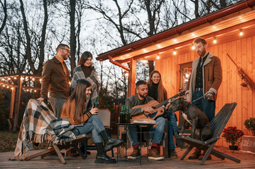 Man playing guitar, dog is sitting on the chair. Group of people is spending time together on the backyard at evening time