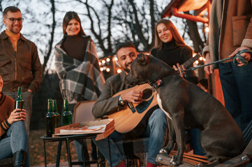 Man playing guitar, dog is sitting on the chair. Group of people is spending time together on the backyard at evening time