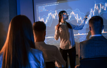 Graphs on projector. Female leader talking to employees in office of stock exchange company