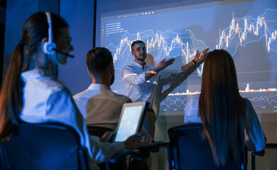 Woman using digital tablet. Male leader talking to employees, showing the plan on the projector in office of stock exchange company