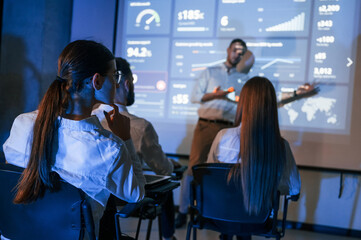 Master class. Male leader talking to employees, showing the plan on the projector in office of stock exchange company