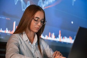 In glasses. Female stock broker is working indoors. Big wide display behind her