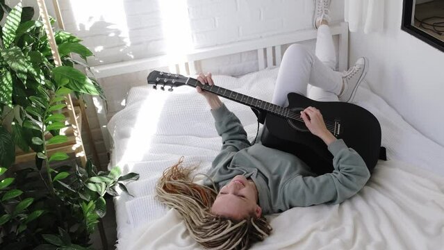 woman with dreadlocks on head singing song and playing acoustic guitar in sunny cottage bedroom, simple living and hobby relaxing