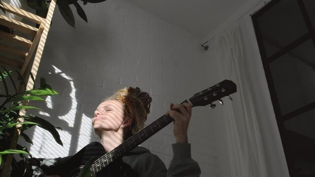 woman with dreadlocks on head singing song and playing acoustic guitar in sunny cottage bedroom, simple living and hobby relaxing