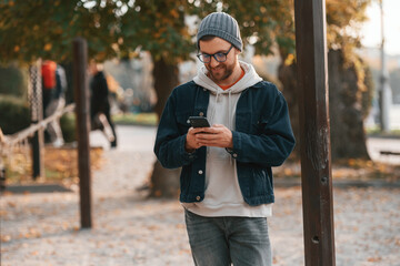 Fototapeta premium Standing with smartphone. Beautiful man in warm clothes is outdoors in the park