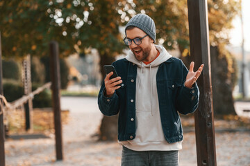 Pleasantly surprised. With smartphone. Beautiful man in warm clothes is outdoors in the park