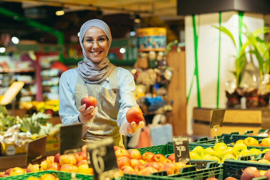 Portrait of saleswoman in supermarket, happy woman in hijab smiling and looking at camera, seller holding apples in vegetable section, Muslim woman in glasses and apron among shelves with goods.
