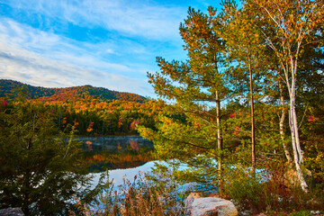 Pine trees and rocks on coast of beautiful lake during peak fall with hills of trees