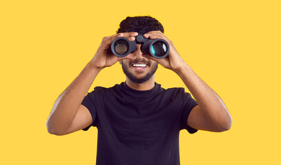Happy ethnic man looking through binoculars looking for profitable advertising offers, isolated on yellow background. Dark-skinned bearded man in black t-shirt looking at you through binoculars