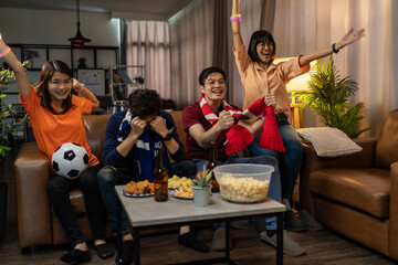 Group of Asian people friends sit on sofa watching and cheering football or soccer games competition on TV together at home.Happy man and woman sport fans celebrating sport team victory sports match  © Yingyaipumi