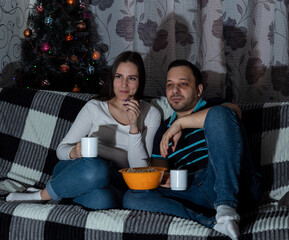 Young woman and man are sitting on couch with mugs, eating popcorn while watching TV. Close-up. Selective focus. Images for articles about movies, hobbies, leisure.
