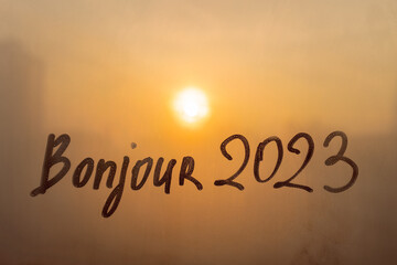 lettering Bonjour in French is hello in english and numbers 2023 paint with finger with streaks of water on splashed by rain foggy glass on orange sunset window © ksenija1803z