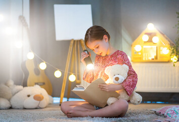 Little beautiful girl reading a book with flashlight in the bedroom. Child reads fairy tale with...