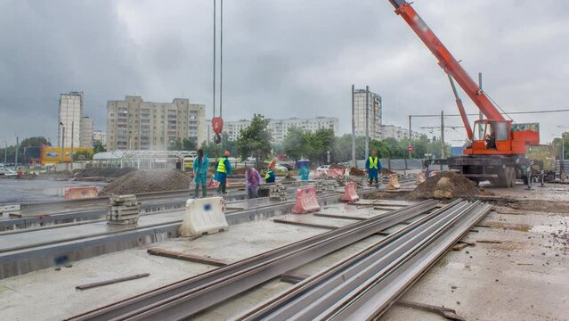 Orange construction telescopic crane unloading tram rails to concrete plates timelapse. Industrial workers with hardhats and uniform. Reconstruction of tram tracks