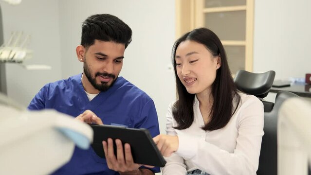 Medicine, dentistry and oral care concept. Confident bearded young man dentist showing tablet pc computer to worried female patient, asian female at modern dental clinic.