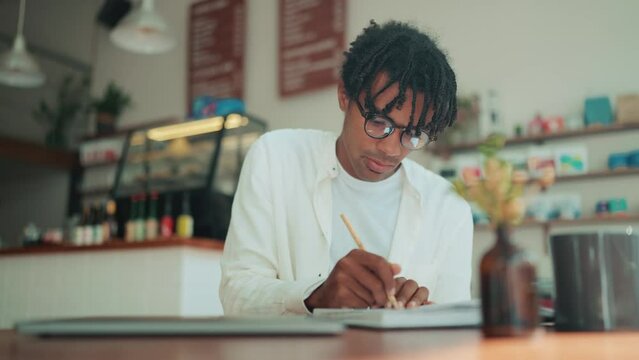 Happy African man with dreadlocks writing something in notepad in the cafe