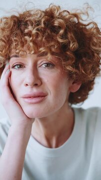 Pensive happy adult curly haired woman looking at camera in the grey studio