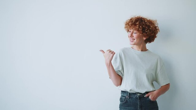 Smiling adult curly haired woman pointing finger to the side in the grey studio
