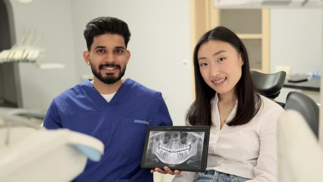 Smiling happy asian woman visiting confident bearded dentist, sitting in dental chair at modern light hospital clinic. Young positive dentist holding tablet pc with x ray scan.