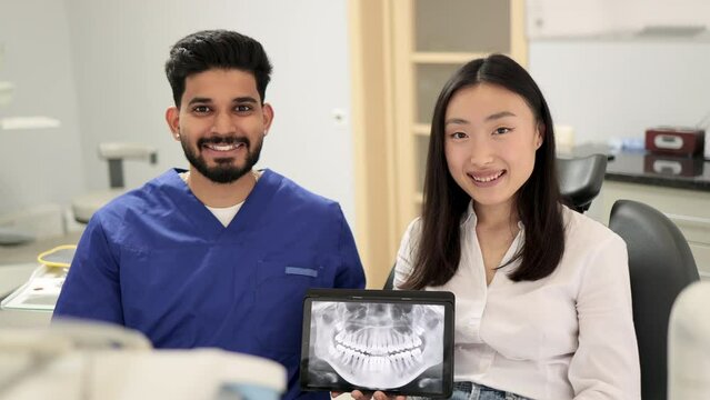 Smiling happy asian woman visiting confident bearded dentist, sitting in dental chair at modern light hospital clinic and showing thumbs up. Young positive dentist holding tablet pc with x ray scan.