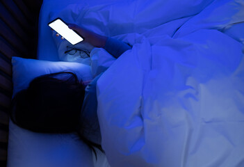 Woman using smartphone lying on bed late at night in bedroom with blue moonlight