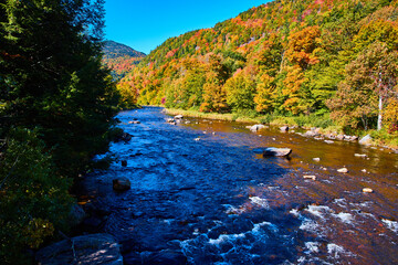 River with small rapids in shade along sunny hills of fall foliage
