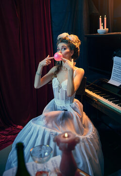 Portrait of young beautiful girl in image of medieval person in elegant white dress sitting at the piano and posing with bubblegum. Comparison of eras, beauty, history, art