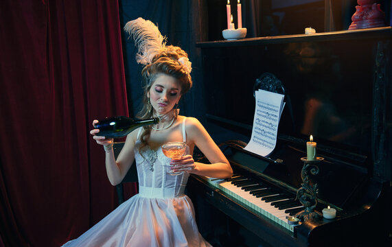 Despair. Portrait of young beautiful girl in image of medieval person in elegant white dress sitting at the piano and drinking wine. Comparison of eras, beauty, history, art