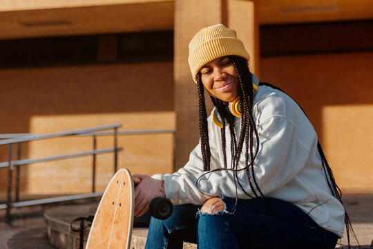 portrait of latin girl with vitiligo, wearing a woolen cap, sitting on the stairs of her high school holding a skateboard. youth culture.