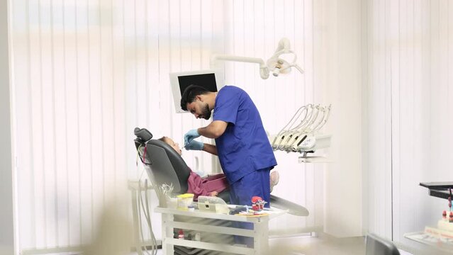 Side view of smiling male professional doctor in blue uniform and gloves, making examination or curing caries and toothache for his little patient, cute kid girl, sitting in dental chair.