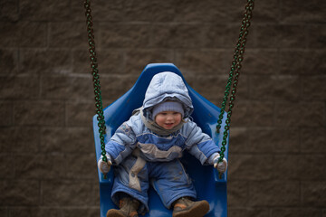chubby Kid with ruddy cheeks rides on a swing in a blue overalls with a hood