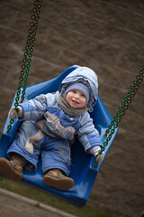 chubby Kid with ruddy cheeks rides on a swing in a blue overalls with a hood