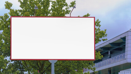 blank advertisement poster with red frame on grey pole near green tree and local city building under cloudy sky