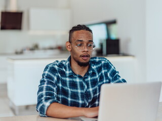 African American man in glasses sitting at a table in a modern living room, using a laptop for business video chat, conversation with friends and entertainment