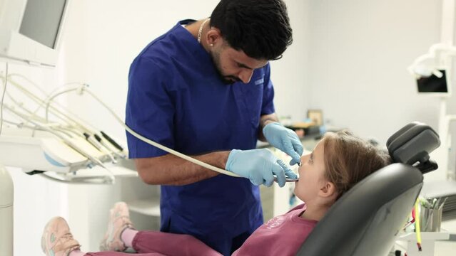 Shot of young confident bearded dentist in blue uniform, making dental check up and caries treatment with drill for her cute little patient, little girl, sitting in dental chair at clinic.