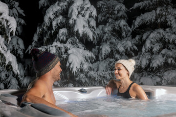 happy couple relaxing in outdoor hot tub at winter with snowy trees in background. spa resort - obrazy, fototapety, plakaty