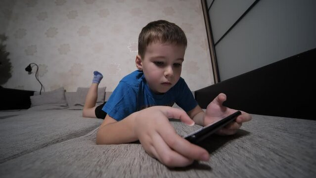 The boy plays on his smartphone while lying on the couch at home.