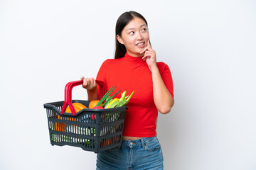 Young Asian woman holding a shopping basket full of food isolated on white background thinking an idea while looking up