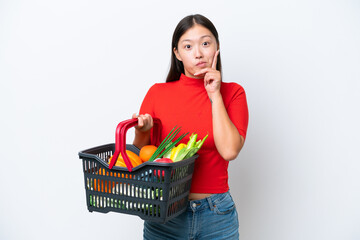 Young Asian woman holding a shopping basket full of food isolated on white background thinking an idea