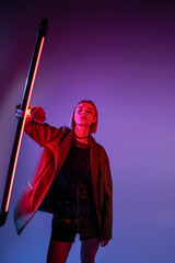 trendy woman in leather jacket and mini skirt posing with glowing neon lamp on purple background.