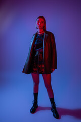 full length of young woman in leather jacket with mini skirt and black boots standing on blue background.