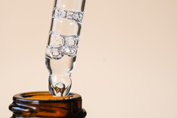 A drop of serum with air bubbles dripping from an eyedropper into a glass vial against a beige background. Side view, macro, space for text.