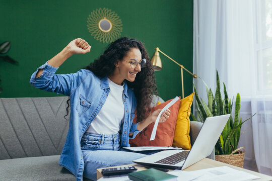 Happy Hispanic Woman At Home Doing Paperwork And Calculating Household Budget, Sitting On Couch In Living Room And Using Laptop At Work, Holding Hand Up Victory And Triumph Gesture.