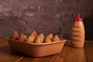 Porção de coxinha sobre mesa de madeira ao lado de pote de pimenta com fundo rústico de tijolos