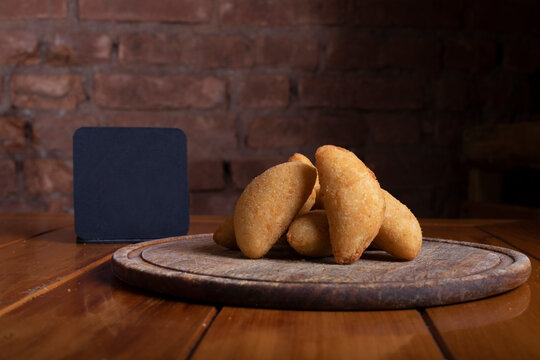 Por&ccedil;&atilde;o de risoles sobre mesa de madeira com placa preta para informa&ccedil;&otilde;es de valor e pre&ccedil;o com fundo r&uacute;stico de tijolos